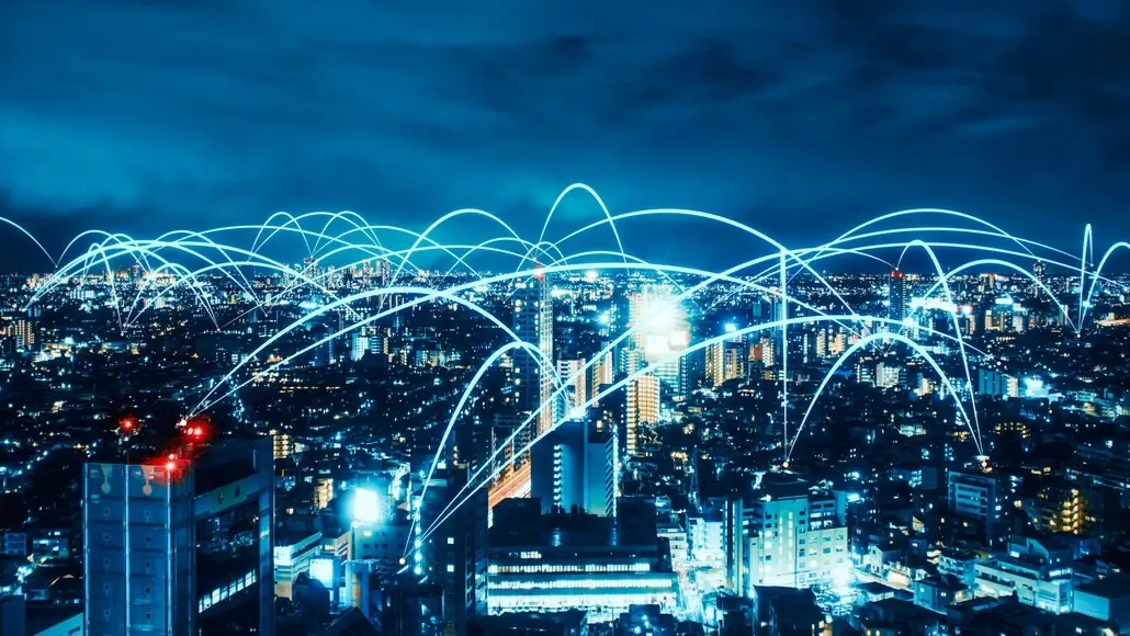 Night cityscape at blue hour with glowing network lines/arcs across the skyline, symbolizing global connectivity and communications.