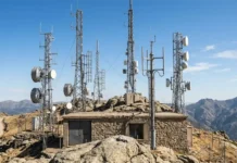 Private ISAC By U.S. Telecom Giants For AI-Led Cyberattacks Communications equipment on a rocky mountaintop with multiple antenna towers and satellite dishes surrounding a stone hut against a blue sky.
