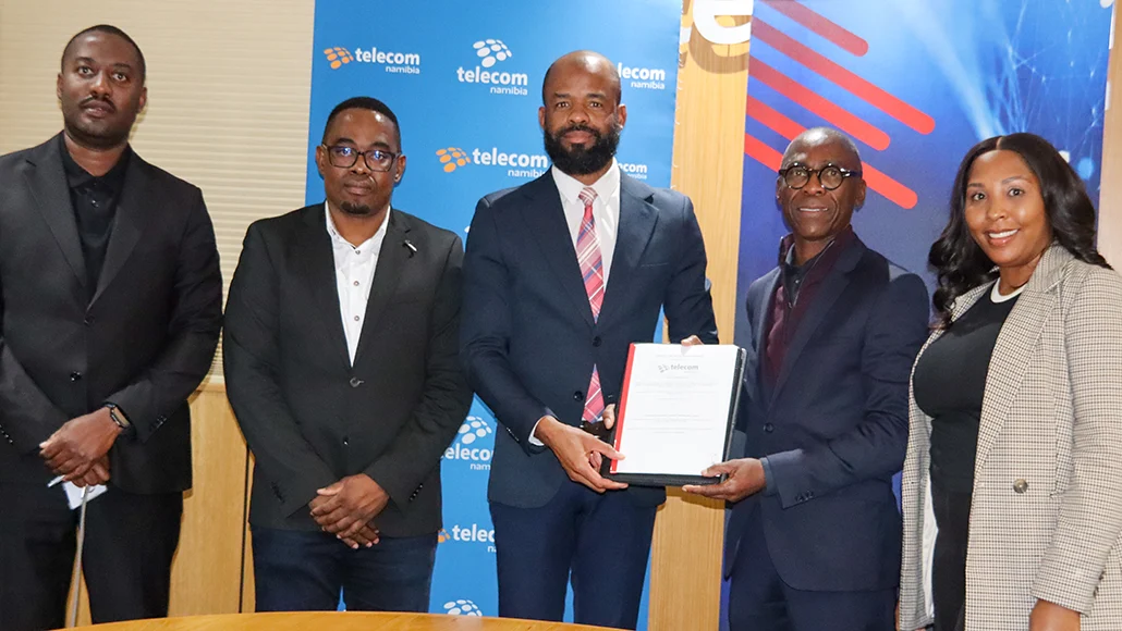 Five professionals in business attire stand together; the two men in the center exchange a certificate in front of a Telecomm Namibia backdrop.