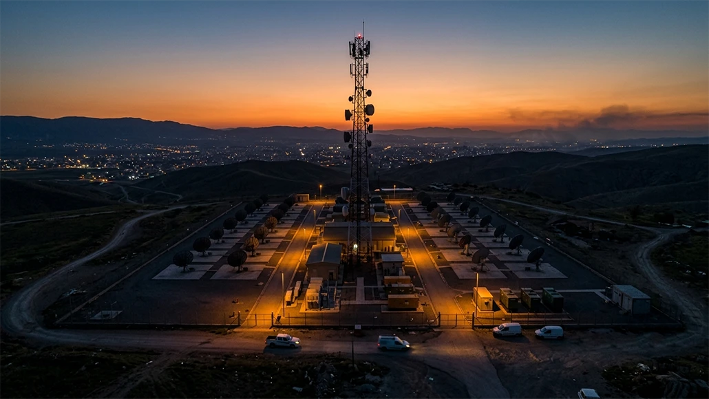 Telecommunications tower surrounded by satellite dishes at a fenced facility on a hill at sunset.