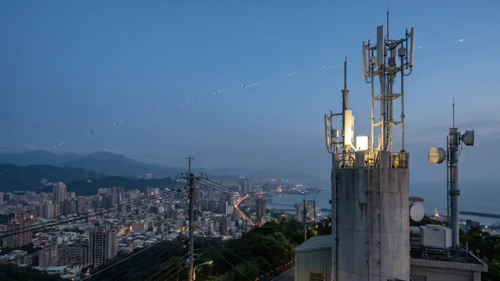 City skyline at dusk with a tall telecom tower and antennas on a hilltop over the city and coastline beyond, including power lines in the foreground.