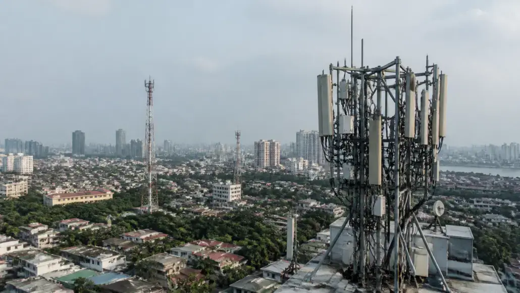 Rooftop cell tower with multiple antennas overlooking a dense urban area and high-rise buildings in the distance.