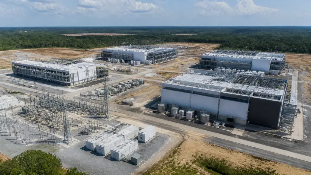 Aerial view of an industrial complex under construction with several large white buildings, electrical substation gear, and storage tanks on barren cleared land near a forest edge.