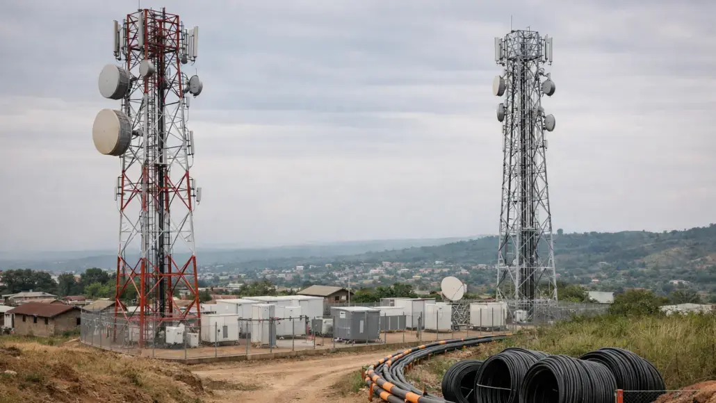Two tall cellular towers with antennas and dishes atop a fenced communications site in a rural valley; coiled cables in the foreground.