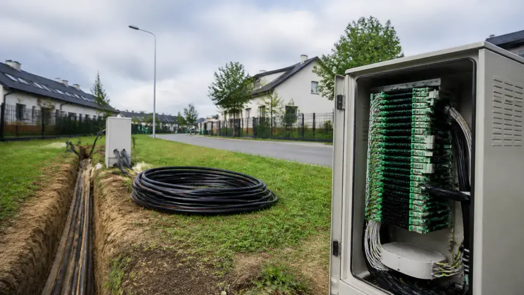Outdoor telecom cabinet with green circuit boards and bundled cables beside a trench in a suburban street visualizing underground fiber deployment.