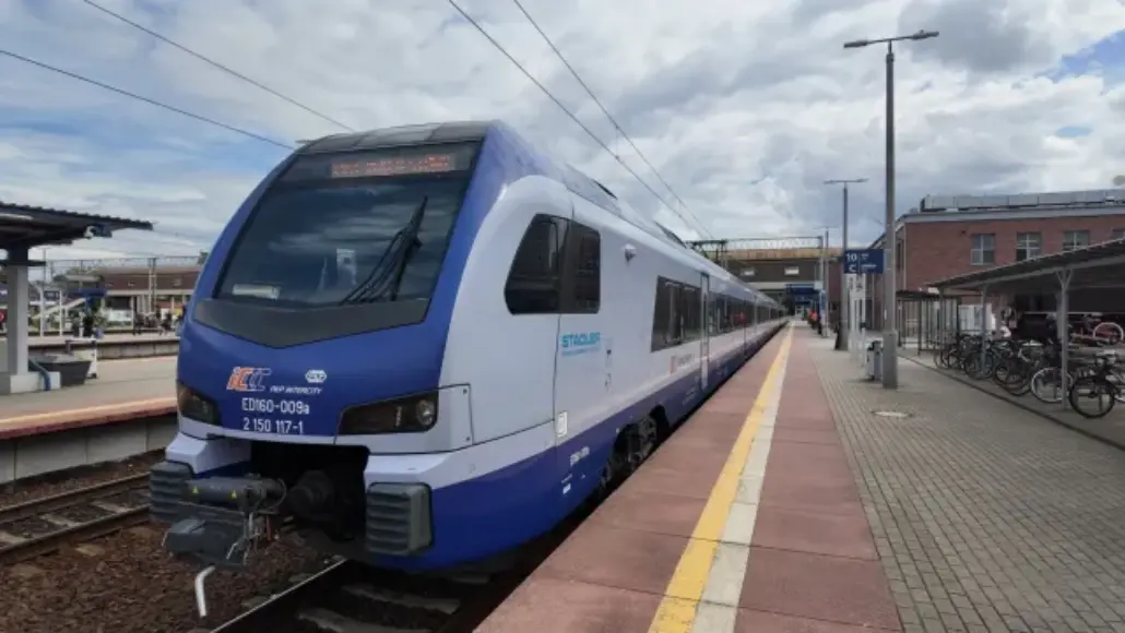 Modern blue-and-white passenger train at a platform, ready for boarding, with bikes and station buildings in the background.