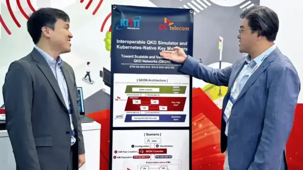 Two men in suits discuss a poster about an interoperable QKD simulator and Kubernetes-native key managers in a conference setting.
