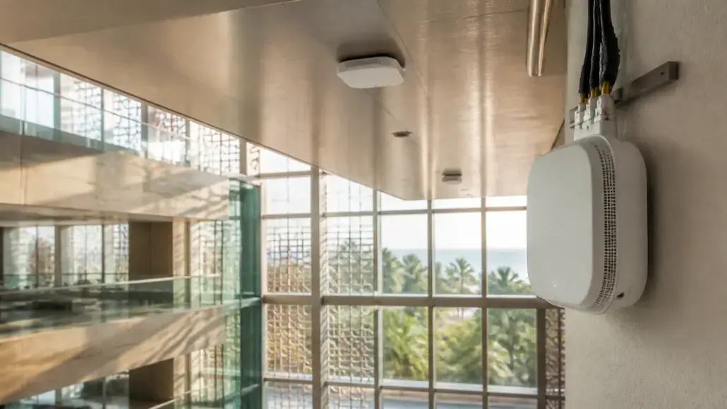 White wall-mounted wireless access point with exposed cables in a bright, modern building, large windows showing palm trees outside.