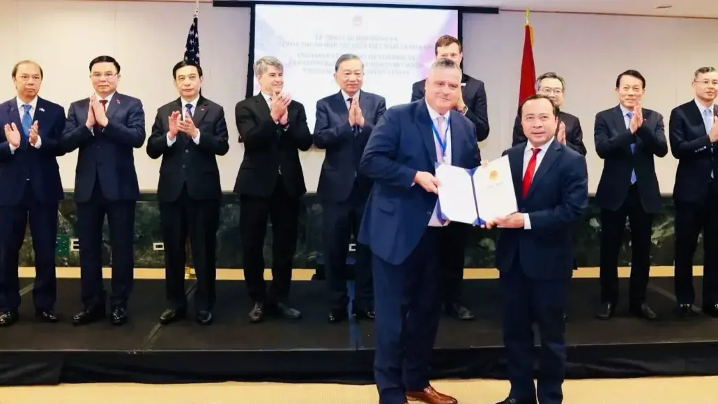 Two men in suits exchange a signed document on a stage as officials applaud in the background.