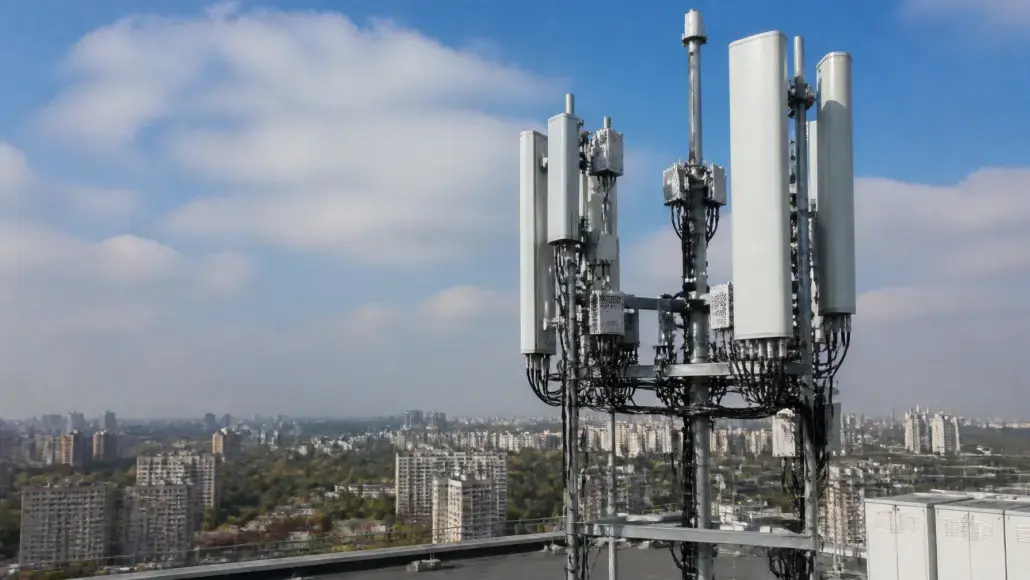 Cellular antenna mast on a rooftop with cables, overlooking a city skyline under a blue sky.